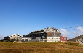 Karl Ludwig Haus, Rax, Schutzhütte, Region Semmering Rax, Wiener Alpen in Niederösterreich, © Wiener Alpen/Bene Croy Panoramablick auf einen Berghütte mit umliegenden Gipfeln unter blauem Himmel.