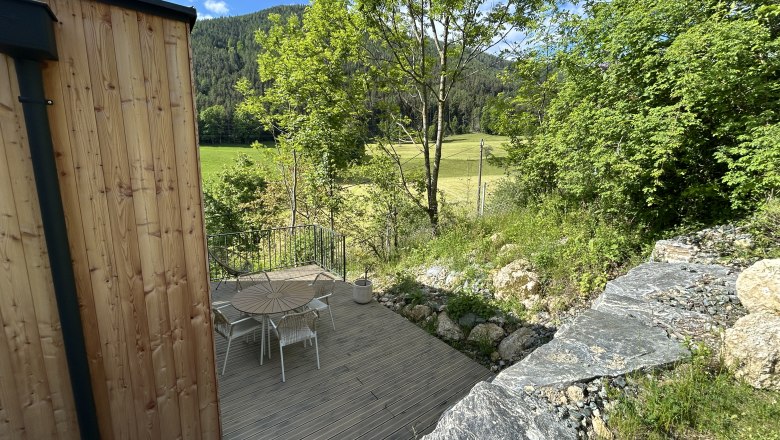 Tiny House, Feigenterrasse am Semmering, © Stupbacher Terrasse mit Holztisch und Stühlen, umgeben von Bäumen und Wiese, mit Blick auf bewaldete Hügel.