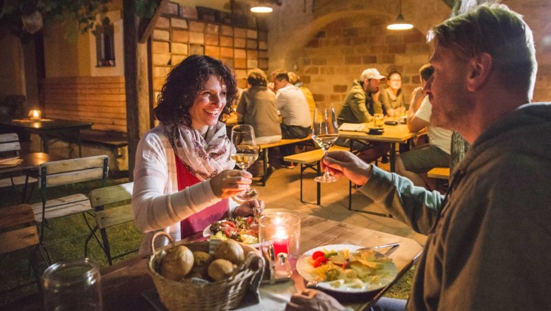 Abendstimmung beim Heurigen, © Daniela Matejschek Ein Paar stößt bei einem Heurigen mit Weingläsern an, im Hintergrund sitzen weitere Gäste an Holztischen.