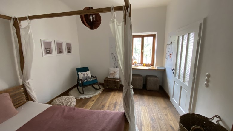 2. Schlafzimmer, © Kathrin Bürger Gemütliches Schlafzimmer mit Himmelbett, Schaukelstuhl und Fensterbank. Holzboden und helle Wände.