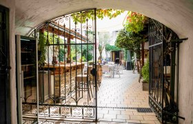 Arkadenhof Maximahl, © Robert Herbst Blick durch ein schmiedeeisernes Tor in einen gemütlichen Innenhof mit Tischen und Pflanzen.