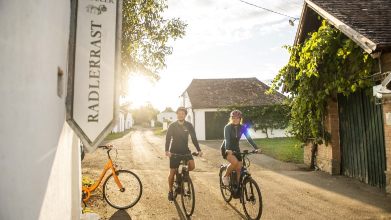 Kellergasse Maulavern, © Weinviertel Tourismus GmbH / POV / Robert Herbst Zwei Radfahrer fahren durch eine sonnige Kellergasse mit Weinkellern und einem Schild mit der Aufschrift 'Radlerrast'.