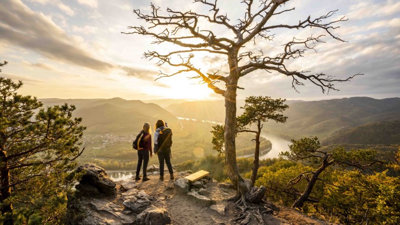 Ausblick von der Kanzel, © Robert Herbst Zwei Personen stehen auf einem Felsen mit Blick auf eine Flusslandschaft bei Sonnenuntergang.