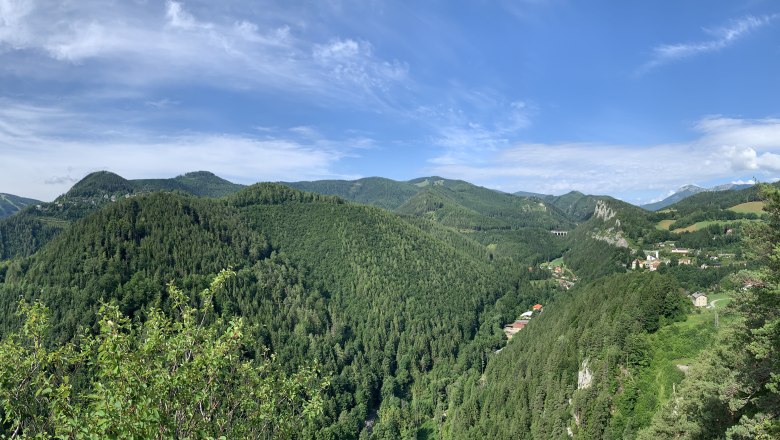 Breitenstein Blick Weinzettlwand, © Tourismusverband Semmering-Rax-Schneeberg Panoramablick auf bewaldete Hügel und ein Tal mit verstreuten Häusern unter blauem Himmel.