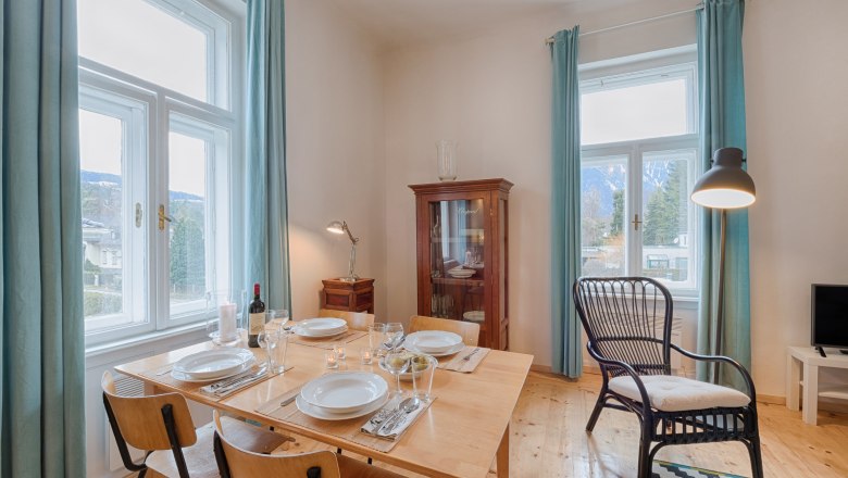 Top 5 - Wohnzimmer, © VP FeWo OG Essbereich in einem Wohnzimmer mit Holztisch, Stühlen, Vitrine und großen Fenstern mit blauen Vorhängen.