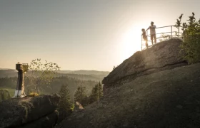 Wachstein in Bad Traunstein, © Studio Kerschbaum Zwei Personen stehen auf einem Felsen mit Geländer, im Hintergrund Sonnenuntergang und Wald.