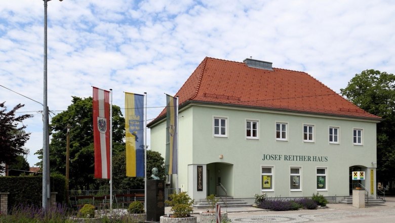 Josef Reither Museum, © Josef Reither Museum - Alois Wurzinger Josef Reither Haus mit Flaggen und Büste im Vordergrund.