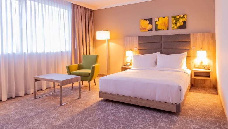 Zimmer, © Hilton Garden Inn Wiener Neustadt Modernes Hotelzimmer mit Doppelbett, grünem Sessel, Tisch und Stehlampe.