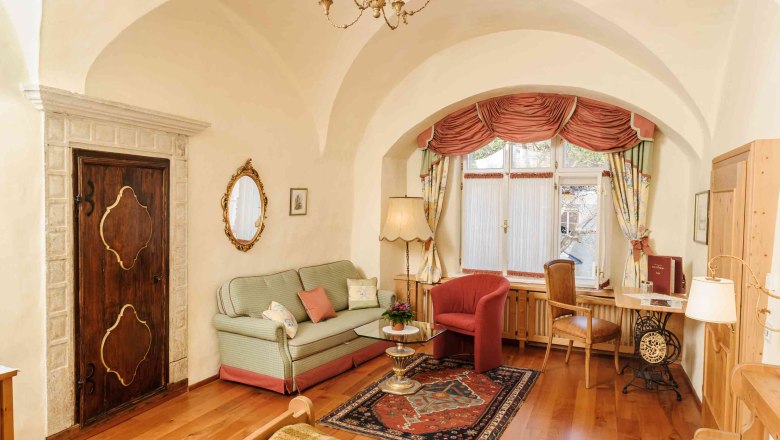 Kirchenwirt Weißenkirchen, © Rita Newman Gemütliches Wohnzimmer mit antikem Flair, Sofa, Teppich, Tisch und Fenster mit Vorhängen.