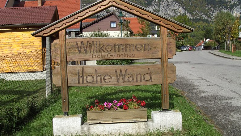Willkommen im Naturpark Hohe Wand, © Stern Holzschild mit der Aufschrift 'Willkommen in der Gemeinde Hohe Wand' vor einer Berglandschaft.
