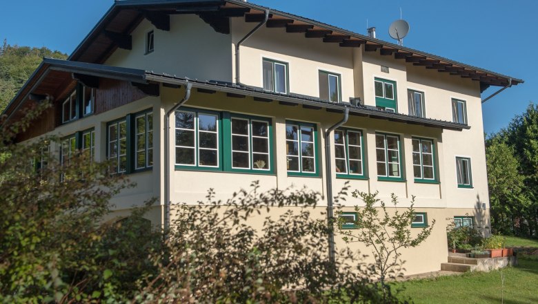 Haus vom Parkplatz aus, © Dorfschmiede eG Ein zweistöckiges Haus mit vielen Fenstern und einem Satteldach, umgeben von Bäumen und Sträuchern, unter einem klaren blauen Himmel.