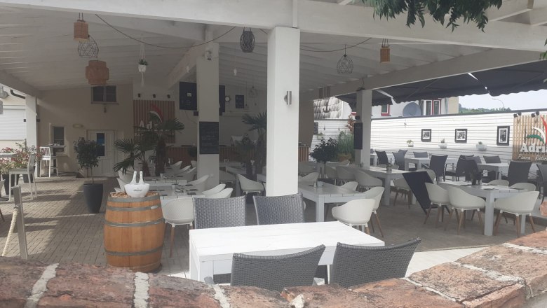 Gemütlicher Gastgarten., © Wiener Alpen Ein überdachter Gastgarten mit weißen Tischen und Stühlen, umgeben von Pflanzen und Dekorationen.