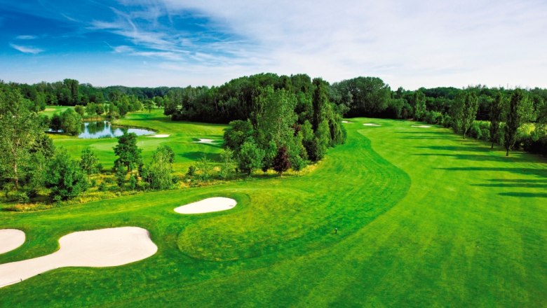 Golf Club Gutenhof, © Golf Club Gutenhof Grüner Golfplatz mit Bunkern und Teich, umgeben von Bäumen unter blauem Himmel.