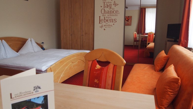 Zimmer, © Bruthenne Ein gemütliches Hotelzimmer mit einem Doppelbett, einem Sofa, einem Tisch und einem Schrank. An der Wand steht ein motivierender Spruch.