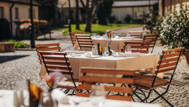 Gastgarten im Innenhof, © Niederösterreich Werbung / Daniela Führer Ein sonniger Gastgarten mit Holztischen und -stühlen, gedeckt mit weißen Tischdecken und Gläsern, in einem gepflasterten Innenhof.