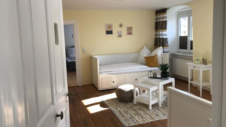 Aufenthaltsraum, © Martina Braun Heller Raum mit gelben Wänden, einem weißen Tagesbett, kleinem Tisch und Pflanzen.