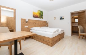 Schlafzimmer, © Schönfelderhof Modernes Schlafzimmer mit Holzmöbeln, Doppelbett, Tisch und Stuhl. Ein Spiegel und Bilder an der Wand, Tür führt zu einem Badezimmer.