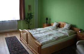 Gästezimmer, © Majewsky Zant Ein gemütliches Gästezimmer mit grün gestrichenen Wänden, einem Holzbett, Nachttischen, einer Pflanze und einem Teppich auf Holzboden.