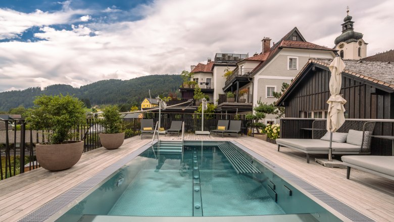 Refugium Lunz, © Niederösterreich Werbung / Maximilian Pawlikowsky Ein moderner Pool auf einer Terrasse mit Blick auf umliegende Häuser und bewaldete Hügel im Hintergrund.