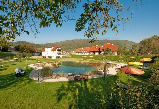 Gartenanlage, © Residenz Wachau Gartenanlage mit Teich, Liegestühlen und Sonnenschirmen, umgeben von Gebäuden und Hügeln.