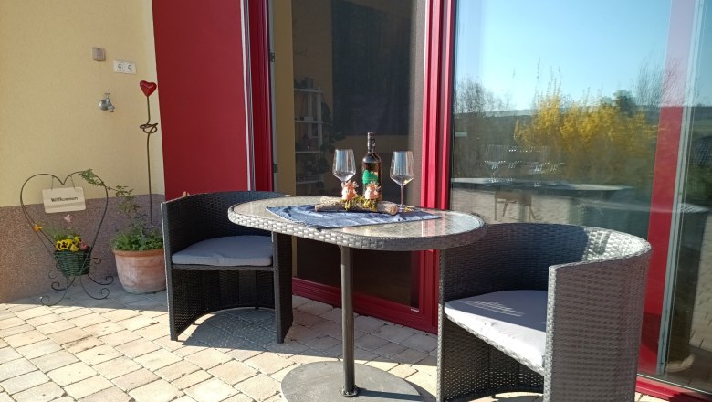 Terrasse, © S. Gritsch-Stockinger Terrasse mit Tisch, zwei Stühlen, Weinflasche und Gläsern vor einer roten Tür.