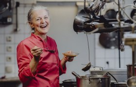 Wirtin Augusta Mayerhofer, © Niederösterreich Werbung/Rita Newman Eine lächelnde Frau in einer roten Kochjacke steht in einer Küche mit einem Löffel und einer Schüssel in der Hand.