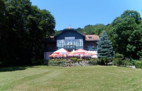 Ansicht Haus, © Rudolfshof Ein großes Haus namens Rudolfshof mit Terrasse und Sonnenschirmen, umgeben von Bäumen und einer Wiese.