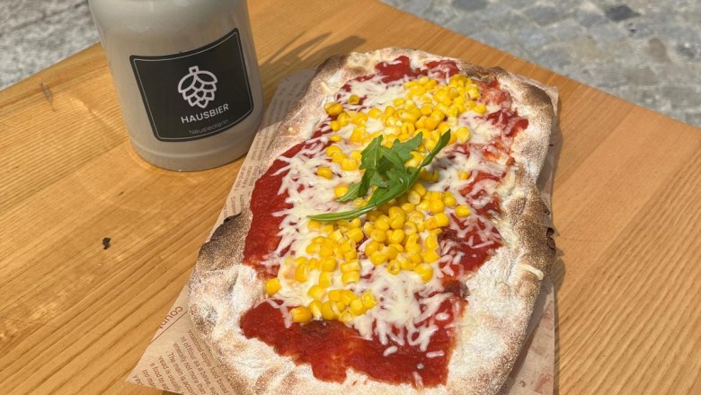 Belegter Fladen, "Die Neusiedlerin", Klein-Neusiedl, © Die Neusiedlerin Fladen mit Mais und Rucola auf einem Holzbrett serviert, daneben ein Behälter mit Besteck und Servietten.