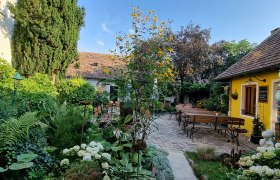 Blick in den Garten, © Fam. Lehninger Ein gemütlicher Garten mit gelbem Gebäude, Holzbänken und üppiger Vegetation unter blauem Himmel.