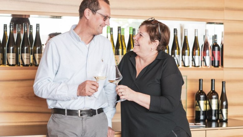 Wolfgang und Elfi Aigner, © Michael Parak Ein Mann und eine Frau stoßen mit Weingläsern an, im Hintergrund Weinflaschen.