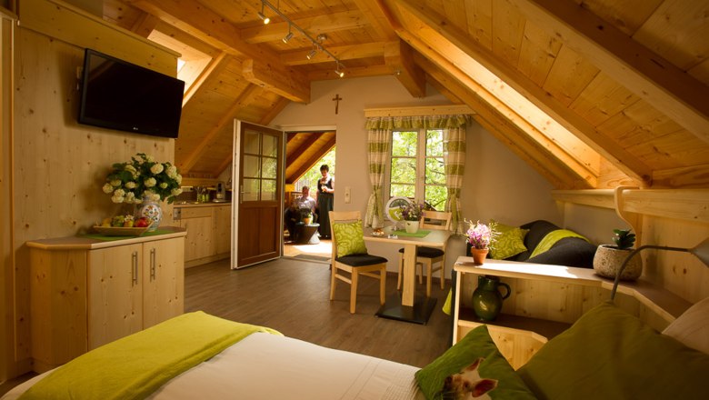 Ferienwohnung beim Bauer und Wirt Langthaler, © Rita Newman Gemütliche Ferienwohnung mit Holzausstattung, Dachschrägen und grünen Akzenten. Im Hintergrund sind zwei Personen auf einem Balkon zu sehen.