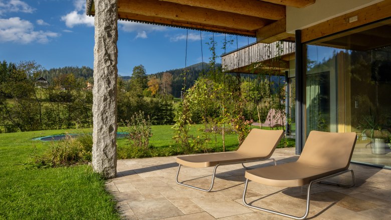 Ferienhaus am See, © Niederösterreich Werbung / Maximilian Pawlikowsky Terrasse eines Ferienhauses mit zwei Liegestühlen, umgeben von grüner Landschaft und Bäumen.