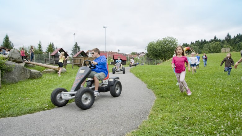 Erlebnisspielplatz, © Pension Bruckner Kinder spielen auf einem Erlebnisspielplatz mit Kettcars und laufen über eine Wiese.