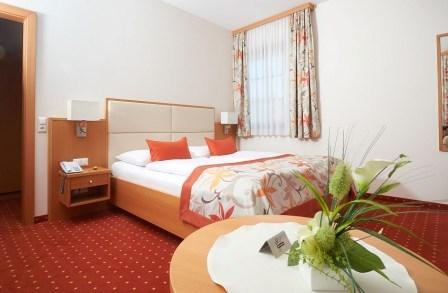 Pension Sprinzl, © Sprinzl Ein gemütliches Hotelzimmer mit Doppelbett, rotem Teppich und Blumendekoration auf einem Tisch.