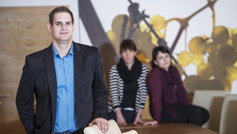 Familie Blaha, © Alfred Kusdat Ein Mann in einem blauen Hemd und schwarzem Sakko steht vor einem Tisch, im Hintergrund sitzen zwei Frauen.