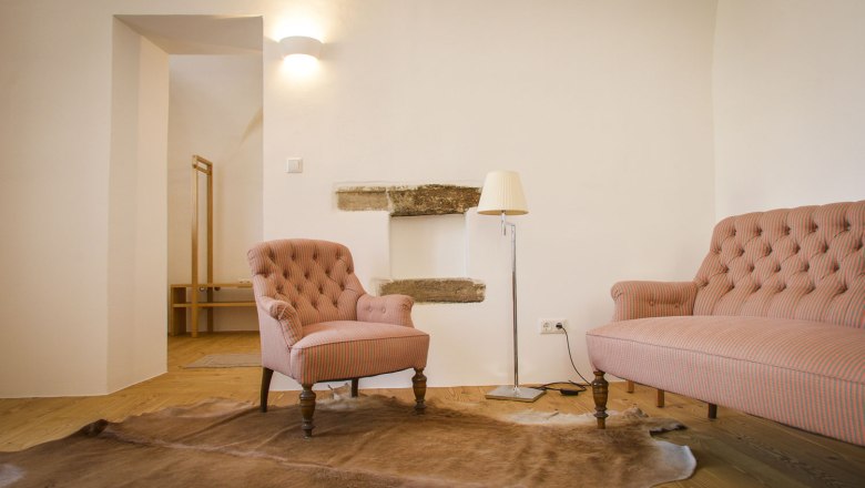 Gut Oberstockstall, © Robert Herbst Ein gemütlicher Raum mit einem rosa gestreiften Sessel und Sofa, einer Stehlampe und einem Fellteppich auf Holzboden.