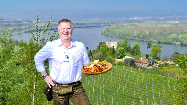 Der Wirt persönlich, © Lumpazi Bräu Ein Mann in traditioneller Kleidung hält ein Tablett mit Essen vor einer Landschaft mit Fluss und Weinbergen.