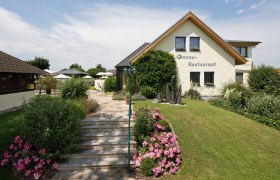 Haus, © DR Ein Restaurant mit Garten und Terrasse, umgeben von Blumen und Büschen.