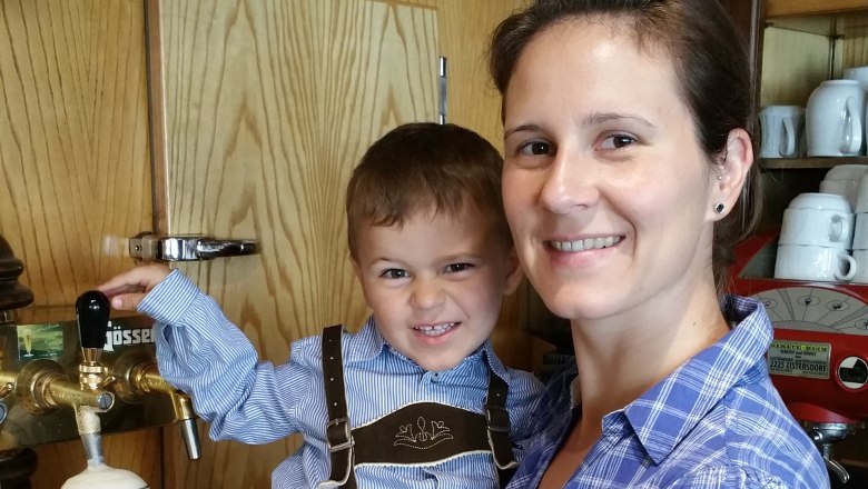 Familie Klapka, © Gasthaus Klapka Eine Frau hält ein Kind in Lederhose an einem Bierzapfhahn. Im Hintergrund sind Spirituosenflaschen zu sehen.