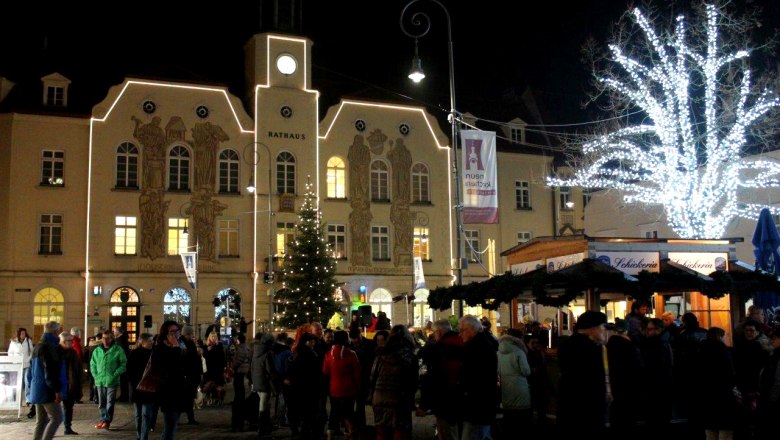 Hauptplatz, © Stadtgemeinde Neunkirchen Nachtaufnahme eines belebten Weihnachtsmarktes vor einem beleuchteten Rathaus mit Weihnachtsbaum und Lichterbaum.
