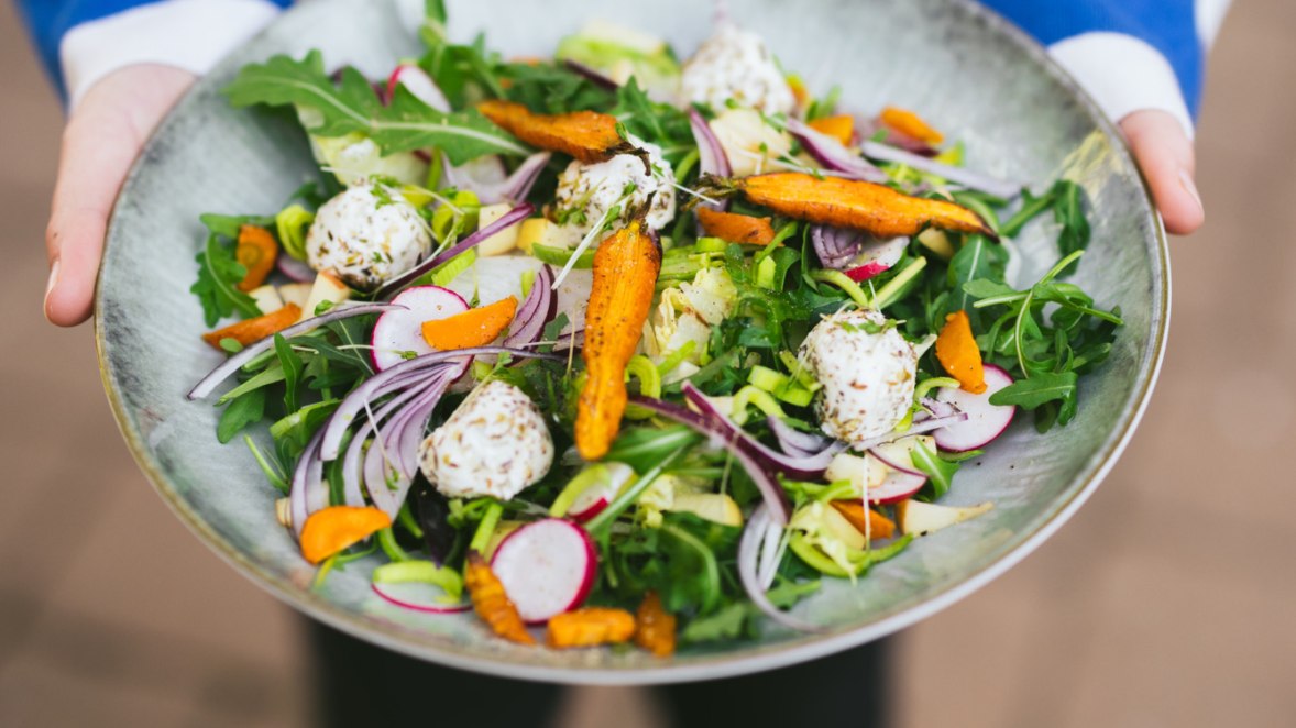Interpretiert klassische Heurigengerichte neu, © Niederösterreich Werbung/Mara Hohla 2 Hände halten Teller mit Ziegenkäsesalat mit Wölblinger Wintersalaten