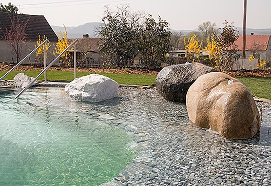 Swimmingpool, © Weinresidenz Sonnleitner Ein Swimmingpool mit großen Steinen und einer Metallleiter, umgeben von Pflanzen und einem Garten.