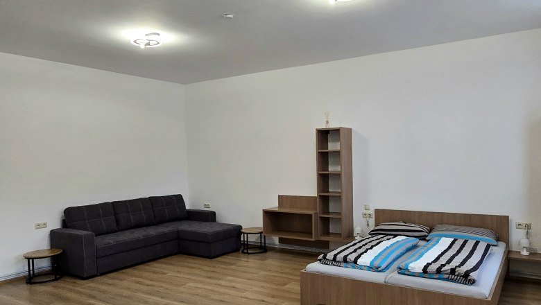 Apartment 3 - Schlafbereich, © Apartment Bauer Zur Alten Schule Ein schlicht eingerichtetes Zimmer mit einem Bett, einem Sofa und einem Regal auf Holzboden.