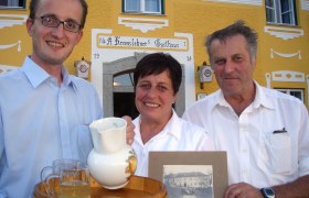 Traditionelle Gastlichkeit, © zVg Drei Personen vor einem gelben Gasthaus mit einem Tablett und einem alten Foto.