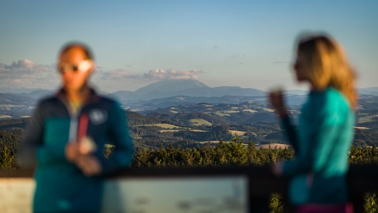 Ausblick von der Aussichtswarte am Hutwisch, © Wiener Alpen/Martin Fülöp Verschwommene Personen vor einer klaren Berglandschaft im Hintergrund. Zu sehen ist der Schneeberg