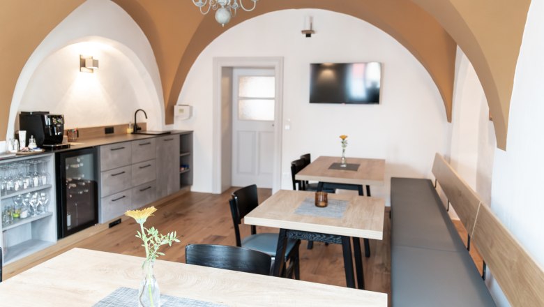 Frühstücks- und Aufenthaltsraum, © Fam. Teufelsbauer Ein gemütlicher Frühstücksraum mit Holztischen, Stühlen und einer Bank. An der Wand hängt ein Fernseher, und es gibt eine Küchenzeile mit Kaffeemaschine.