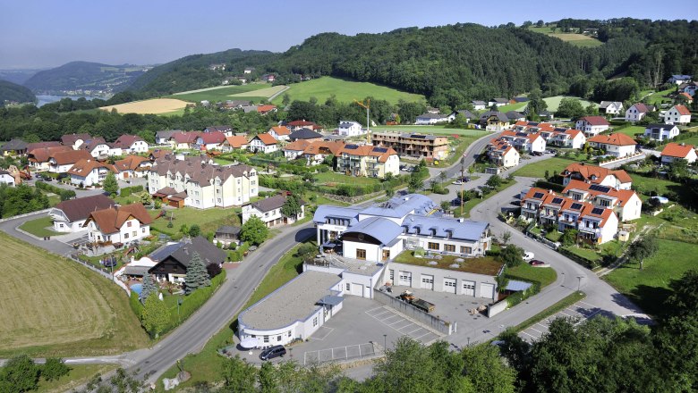 Gemeindezentrum, © Gemeinde Luftaufnahme eines Dorfes mit Gemeindezentrum und umliegenden Häusern in einer grünen Hügellandschaft.