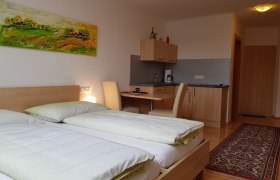 Zimmer, © BIO-Weingut Poys Ein modernes Zimmer mit zwei Betten, einem Schreibtisch, einer kleinen Küchenzeile und einem bunten Gemälde an der Wand.