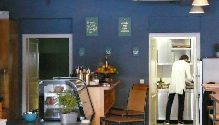 die Theke, © Süd - das Kulturcafé Innenansicht eines Cafés mit blauer Wand, Theke, Barhockern und einer Person in der Küche.