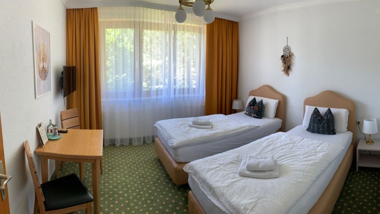 Doppelzimmer Einzelbetten, © Dorfschmiede eG Ein Hotelzimmer mit zwei Einzelbetten, einem Tisch mit Stühlen und einem Fenster mit Vorhängen.